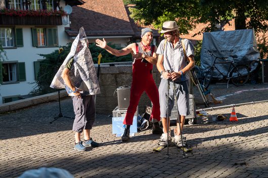Drei Strassenkünstler auf einem Kopfsteinpflasterplatz: Eine Person trägt eine kegelförmige Papiermaske über dem Kopf, während eine zweite in rotem Overall gestikuliert und eine dritte mit Wanderstöcken zuschaut.