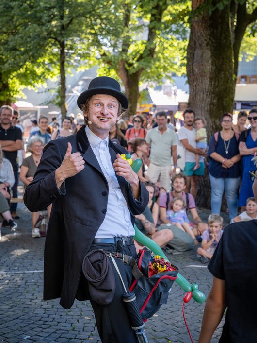 Ein Strassenkünstler in schwarzem Frack und Melone formt bunte Luftballons und zeigt einen Daumen hoch, während eine grosse Menschenmenge ihm auf einem sommerlichen Stadtfest zuschaut.