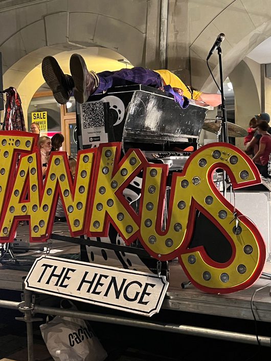 Eine Bühne mit einem grossen gelb-roten Leuchtbuchstaben-Schild mit der Aufschrift 'Tankus the Henge', während eine Person in bunter Kleidung kopfüber über ein schwarzes Keyboard gebeugt liegt. Im Hintergrund sind Zuschauer und ein Mikrofon auf einem Ständer zu sehen.