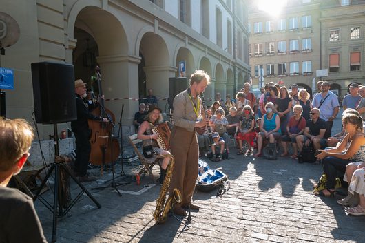 Ein Musiker spielt Klarinette auf einem belebten Stadtplatz in Bern, während eine grosse Menschenmenge dem Strassenkonzert zuschaut. Im Hintergrund sind weitere Bandmitglieder mit Kontrabass und Akkordeon sowie historische Laubengänge zu sehen.