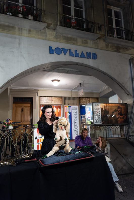 Eine Frau in schwarzem Kleid führt nachts vor einem Geschäft namens «Loveland» eine Puppenshow mit einer provokanten Handpuppe auf. Im Hintergrund sitzen zwei Zuschauer und beobachten die Darbietung.