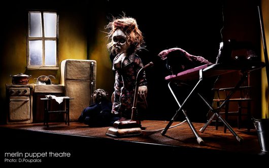Eine detaillierte Puppentheater-Szene zeigt zwei Marionetten in einer düsteren Küchenszenerie, wobei eine stehende Puppe mit zerzaustem Haar über einer am Boden liegenden Puppe steht. Die dramatisch beleuchtete Miniaturkulisse enthält einen Herd, einen Kühlschrank, ein Bügelbrett und einen Staubsauger.