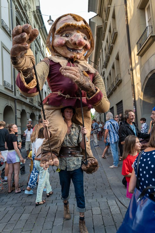 Ein Strassenkünstler trägt eine riesige Gnomen-Marionette auf den Schultern und führt sie durch eine belebte Altstadtgasse, während Zuschauer aller Altersgruppen zuschauen.
