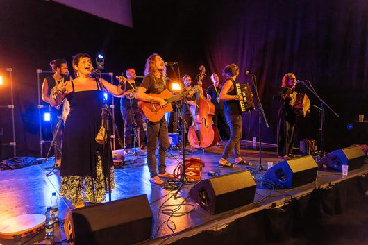 Eine Musikgruppe mit etwa acht Personen tritt auf einer Bühne auf, darunter eine Sängerin im schwarzen Kleid, ein Gitarrist, ein Kontrabassist und zwei Akkordeonspielende.
