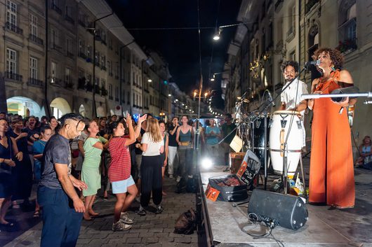 Eine Sängerin im orangefarbenen Overall und ein Musiker treten nachts auf einer Freilichtbühne in einer belebten europäischen Altstadt auf, während das Publikum vor der Bühne tanzt und feiert.