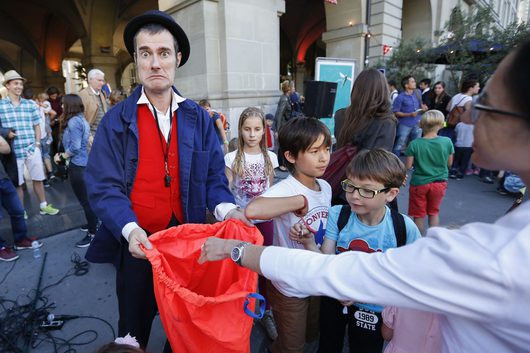 Ein kostümierter Strassenkünstler mit schwarzem Hut und blau-rotem Kostüm hält einen roten Stoffbeutel, während Kinder und Erwachsene bei einer Aussenveranstaltung um ihn herumstehen. Der Künstler macht einen übertriebenen, komischen Gesichtsausdruck in die Kamera.