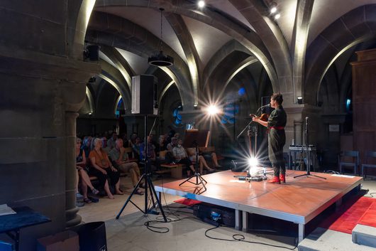 Eine Musikerin spielt Gitarre und singt auf einer kleinen Bühne in einem mittelalterlichen Gewölbekeller vor einem sitzenden Publikum. Die Veranstaltung findet in einem historischen Steingebäude mit gotischen Kreuzgewölben statt.