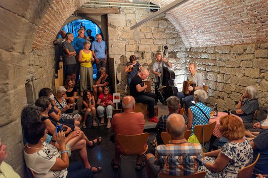 Ein kleines Musikensemble spielt in einem historischen Gewölbekeller vor einem dicht gedrängten Publikum. Drei Musiker mit Kontrabass, Mandoline und Akkordeon treten in einem Raum mit Backsteinmauern auf, während Zuschauer von allen Seiten zuhören.