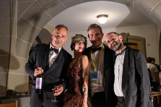 Vier Personen posieren gemeinsam für ein Foto in einem gewölbten Innenraum bei einer festlichen Veranstaltung. Eine Frau in einem aufwendigen Kostüm mit Kopfschmuck steht zwischen drei Männern in Abendgarderobe.