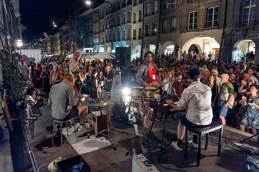 Eine Band spielt bei Nacht auf einer Freiluftbühne in einer belebten Altstadt-Gasse vor einer grossen Menschenmenge. Mehrere Musiker mit Gitarren, Bongos und Keyboard sind auf der Bühne zu sehen, während das Publikum dicht gedrängt zuschaut.