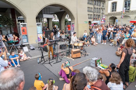 Eine Sängerin und ein Schlagzeuger treten bei einem Strassenmusikfestival in der Berner Altstadt auf, umgeben von einer grossen Zuschauermenge. Die Musikerinnen und Musiker spielen auf einer abgesperrten Strasse unter historischen Laubenbögen.