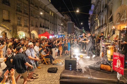 Eine Band spielt nachts auf einer belebten Strassenkulisse in der Berner Altstadt, während eine grosse begeisterte Menschenmenge tanzt und applaudiert. Ein Schild mit der Aufschrift «Buskers Bern» ist rechts im Bild sichtbar.