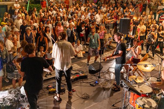 Eine Band spielt auf einer kleinen Bühne im Freien vor einer grossen, dicht gedrängten Menschenmenge bei einem Strassenfestival. Mehrere Musiker mit Gitarre, Geige und Schlagzeug sind von hinten zu sehen, während das Publikum begeistert zuschaut.