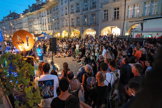 Eine grosse Menschenmenge versammelt sich auf einem gepflasterten Stadtplatz bei Abenddämmerung und beobachtet eine Strassenperformance mit Feuer und Lautsprechern vor historischen Laubengebäuden.