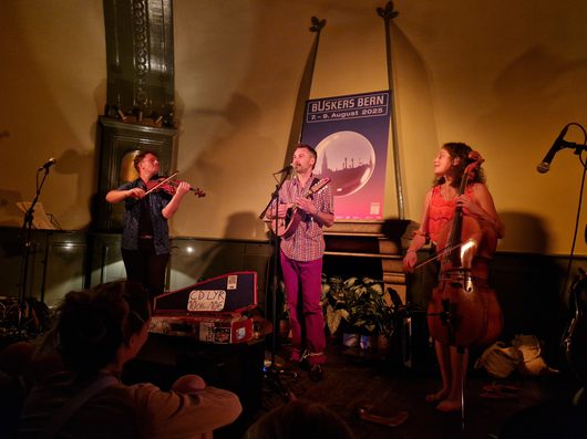 Ein Trio aus Geiger, Gitarrist und Cellistin tritt in einem warmbeleuchteten Innenraum beim Buskers Bern Festival 2025 auf. Im Hintergrund ist ein Festivalplakat mit dem Datum 7.–9. August 2025 sichtbar.