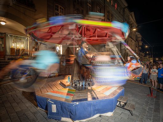 Ein handgefertigtes Kettenkarussell dreht sich bei Nacht auf einem Stadtplatz, während ein Mann in der Mitte die Konstruktion bedient und Zuschauer am Rand zuschauen. Die Langzeitbelichtung erzeugt farbige Bewegungsstreifen der rotierenden Sitze.