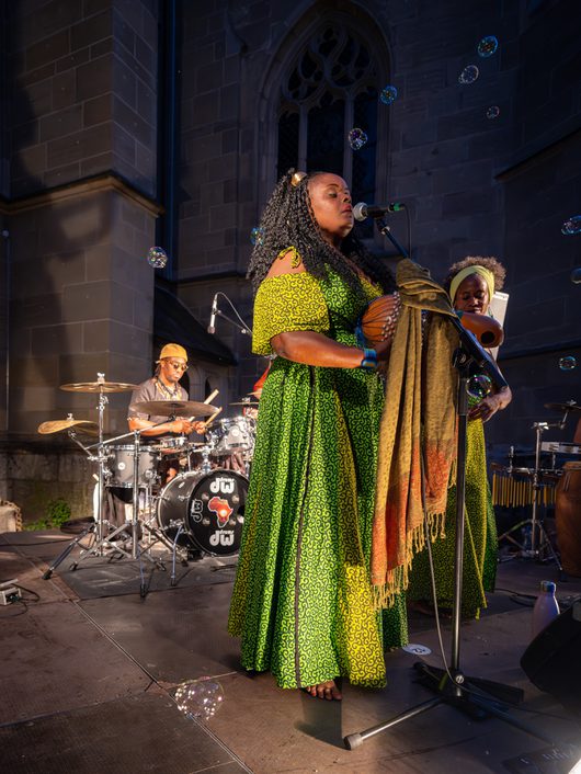 Eine Sängerin in einem leuchtend grünen afrikanischen Kleid singt an einem Mikrofon auf einer Freiluftbühne vor einer Kirche, während Seifenblasen durch die Luft schweben. Im Hintergrund spielen ein Schlagzeuger und eine weitere Musikerin auf der Bühne.