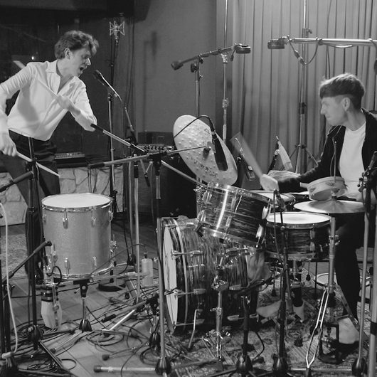 Zwei Musiker spielen in einem Aufnahmestudio an einem grossen Schlagzeug-Setup, wobei einer singt und der andere die Drums schlägt. Das Schwarz-Weiss-Foto zeigt die energiegeladene Atmosphäre einer Live-Session.