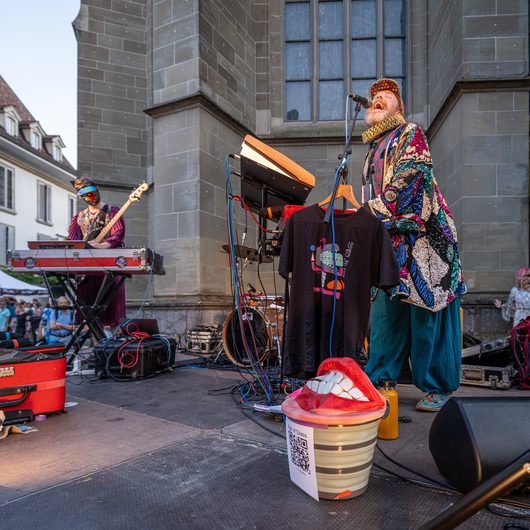 Ein bärtiger Sänger in einem bunten Paillettenmantel und gemusterter Mütze performt lautstark an einem Mikrofon auf einer Freilichtbühne vor einer historischen Steinkirche. Im Hintergrund spielt ein weiterer Musiker Keyboard, während auf der Bühne ein dekoratives Lippen-Requisit und schwarze Band-T-Shirts auf einem Kleiderbügel zu sehen sind.