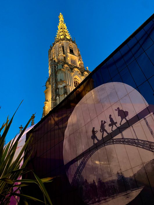 Das beleuchtete Berner Münster ragt vor einem tiefblauen Abendhimmel auf, während im Vordergrund eine Fassade mit einem Kunstmotiv von Musikersilhouetten auf einer Brücke zu sehen ist. Im unteren Bildbereich sind grüne Pflanzenblätter sichtbar.