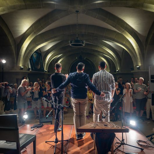 Drei Musiker stehen mit dem Rücken zur Kamera auf einer Bühne in einem gewölbten Saal und verbeugen sich vor einem applaudierenden Publikum. Im Vordergrund ist ein Zupfinstrument auf einem Ständer zu sehen.