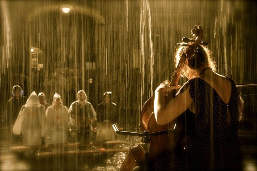 Eine Cellistin spielt im strömenden Regen, während eine Gruppe von Zuschauern in Regenponchos ihr von vorne zuschaut. Die Szene ist in warmes, goldenes Licht getaucht.