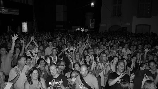 Eine grosse, begeisterte Menschenmenge feiert bei einem Strassenfest oder Open-Air-Konzert bei Nacht. Viele Personen heben die Arme, klatschen und jubeln in die Kamera.