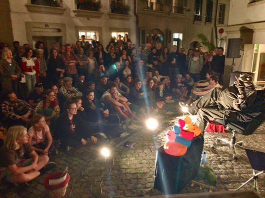 Eine grosse Menschenmenge verfolgt bei Nacht auf einem gepflasterten Platz eine Strassentheatervorstellung mit bunten Figuren und einem kostümierten Performer. Die Zuschauer sitzen und stehen dicht gedrängt vor historischen Gebäuden.