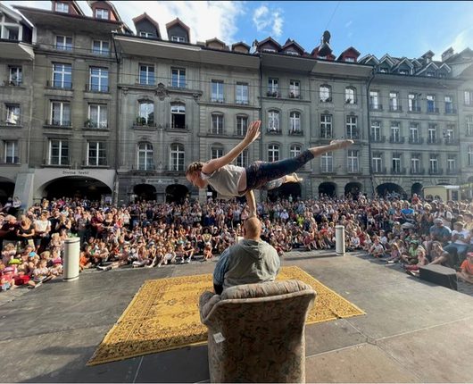 Ein Akrobat wird von einem sitzenden Partner auf einem Sessel in die Luft gehoben und balanciert waagrecht auf dessen Füssen, vor einer grossen Zuschauermenge auf einem Stadtplatz.