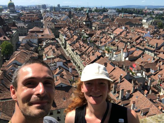 Ein Mann und eine Frau machen ein Selfie auf einem Aussichtspunkt hoch über der Berner Altstadt mit ihren charakteristischen roten Ziegeldächern und historischen Türmen im Hintergrund.