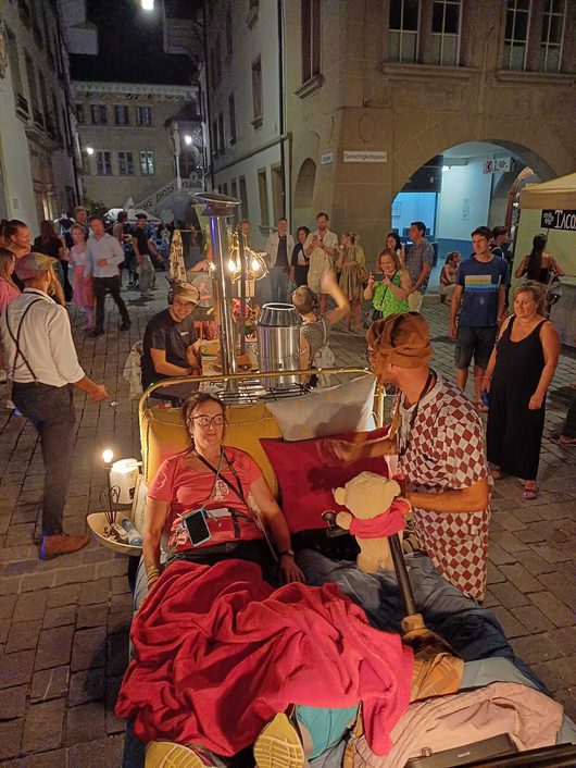 Eine Frau liegt auf einem dekorierten Bett auf Rädern in einer belebten Altstadtgasse bei Nacht, während ein Mann in kariertem Hemd neben ihr sitzt und Zuschauer das Strassentheater beobachten.