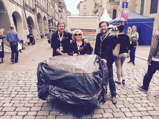 Drei Personen posieren lächelnd hinter einem grossen, mit einer dunklen Plane abgedeckten Gepäckstück auf einem Kopfsteinpflasterplatz beim Buskers Bern Street Music Festival. Im Hintergrund sind historische Laubengänge, ein blaues Festzelt und ein Festivalschild sichtbar.