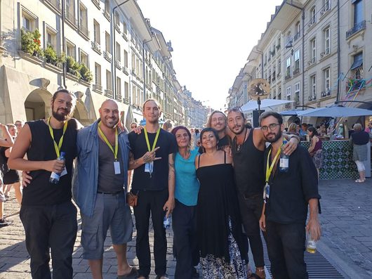 Eine Gruppe von neun lächelnden Personen posiert gemeinsam auf einer belebten Fussgängerzone in einer europäischen Altstadt bei strahlendem Sonnenschein. Mehrere Personen tragen gelb-grüne Akkreditierungsbänder, was auf eine Veranstaltung oder ein Festival hindeutet.