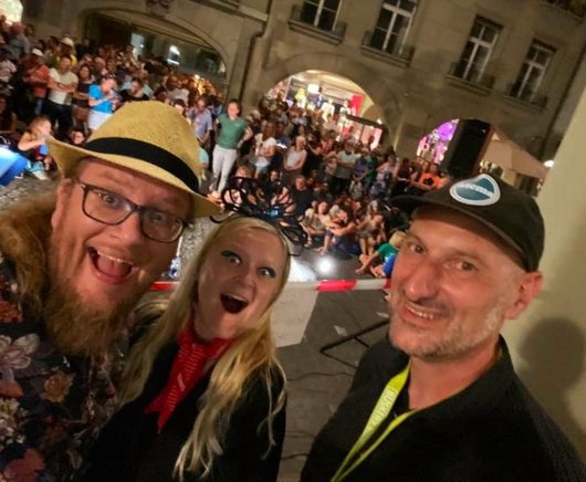 Drei lachende Personen machen ein Selfie vor einer grossen Menschenmenge bei einem nächtlichen Strassenfest in einer Altstadt. Im Hintergrund sind Hunderte von Zuschauern auf einem belebten Platz zu sehen.