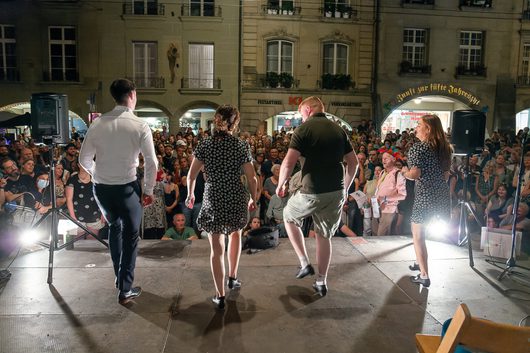 Vier Tänzerinnen und Tänzer treten auf einer beleuchteten Freiluftbühne vor einem grossen Publikum auf einem Stadtplatz auf. Die Aufführung findet vor einem historischen Gebäude mit Laubengang auf einem abendlichen Strassenfest statt.