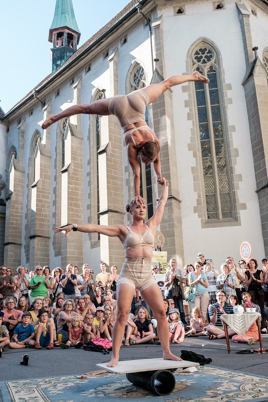 Zwei Akrobatinnen führen eine Gleichgewichtsbalance-Nummer auf einem Rollbrett vor einer gotischen Kirche auf, wobei eine Performerin auf den Händen der anderen steht und ihre Beine in die Luft streckt. Eine grosse Menschenmenge schaut der Strassenshow im Freien zu.