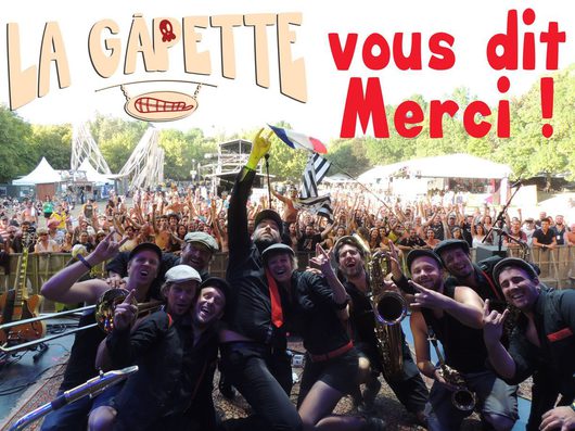 Eine Musikband posiert jubelnd auf einer Freilichtbühne vor einer grossen, begeisterten Festivalmenge. Im Vordergrund halten Bandmitglieder Instrumente wie ein Saxofon und eine Gitarre, während im Hintergrund die Zuschauer die Hände in die Luft strecken.