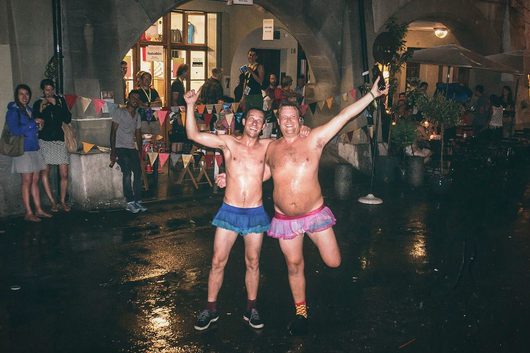 Zwei oberkörperfreie Männer in bunten Tüllröcken posieren lachend und mit ausgestreckten Armen auf einem nassen Platz bei Nacht. Im Hintergrund sind Zuschauer, bunte Wimpelketten und ein beleuchtetes Festivalbüro unter historischen Bögen zu sehen.