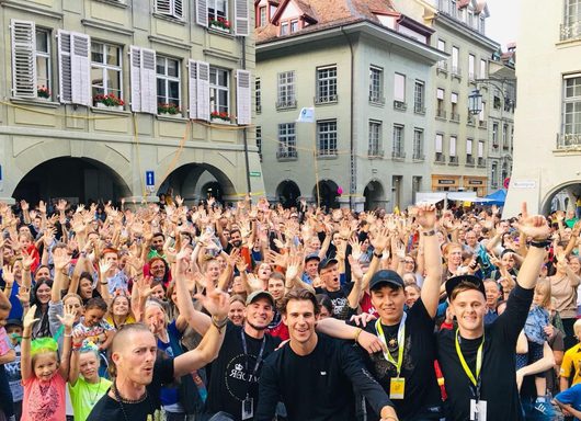 Eine grosse, fröhliche Menschenmenge hebt die Hände auf einem historischen Stadtplatz, während vier junge Männer mit Akkreditierungsbändern im Vordergrund lächeln und jubeln. Im Hintergrund sind klassische mitteleuropäische Altstadtgebäude mit Fensterläden und Blumenkästen zu sehen.
