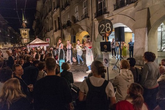 Mehrere Sängerinnen treten bei einem nächtlichen Strassenfest in einer historischen Altstadt vor einer grossen Menschenmenge auf. Im Hintergrund ist ein beleuchteter Uhrturm und ein Laubbogen mit der Nummer 28 zu sehen.