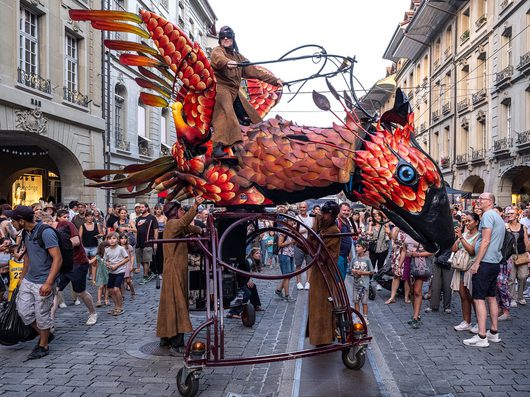 Eine riesige mechanische Fisch-Skulptur mit leuchtend roten und orangefarbenen Schuppen wird von Performern in braunen Kutten durch eine belebte Fussgängerzone gefahren, während eine Person in Fliegerbrille auf dem Rücken des Wesens steht. Eine grosse Menschenmenge säumt die Strasse und beobachtet die Strassentheater-Aufführung in der Berner Altstadt.